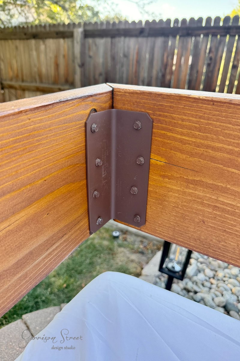 Close-up of two wooden 2x8s joined at a corner with a brown metal bracket and several screws. In the background, there is a fence, rocks, grass, and some blurred outdoor elements.