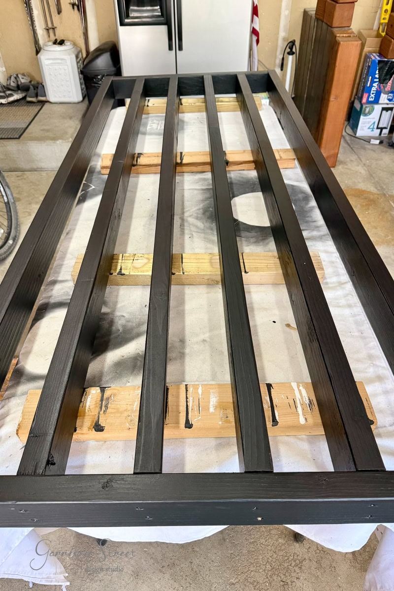 A black wooden bed frame with four horizontal slats is drying on a tarp-covered table, supported by wooden planks, with paint marks visible on the tarp.