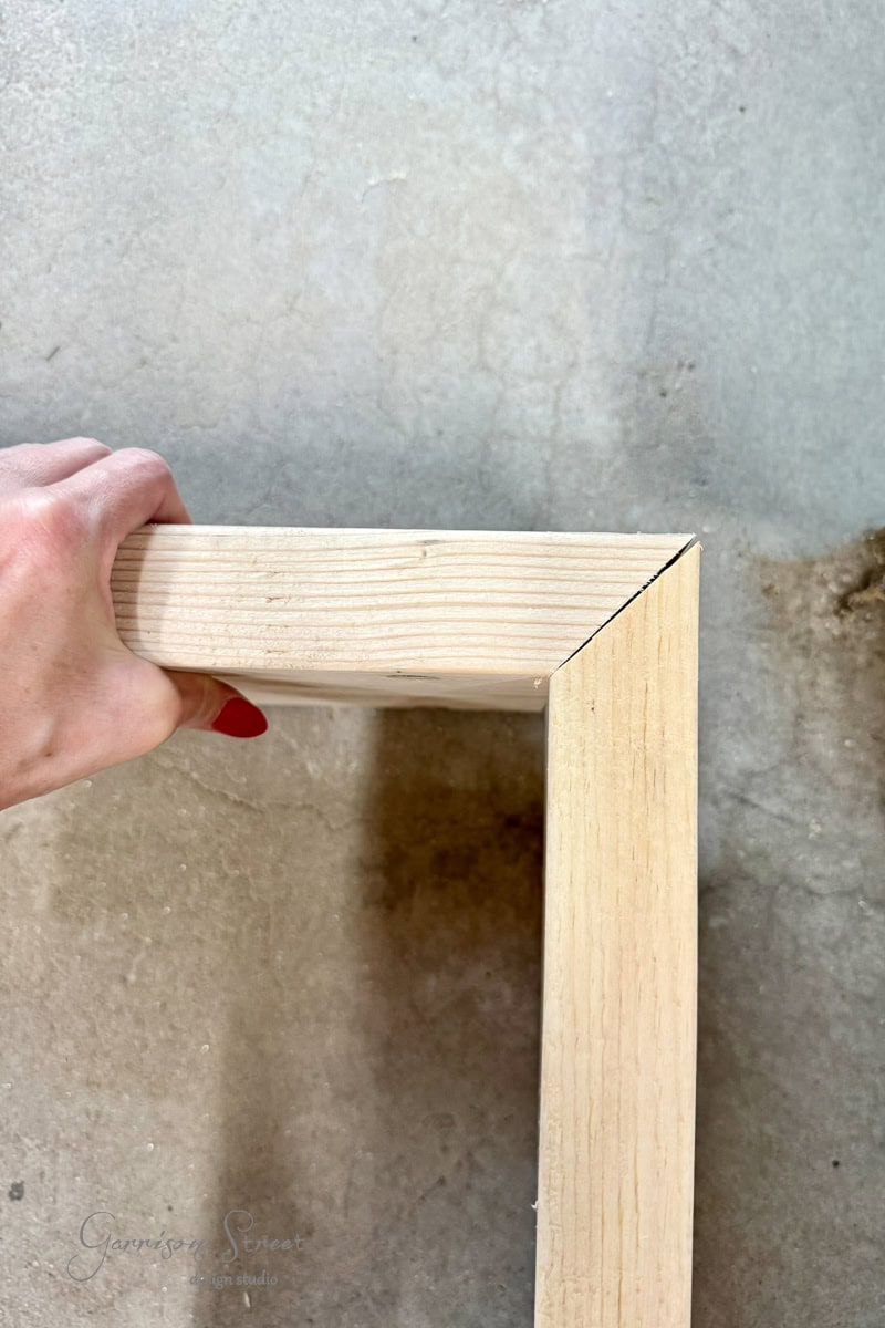 A hand holds the corner of an unfinished wooden frame with a mitered joint, resting on a concrete surface.
