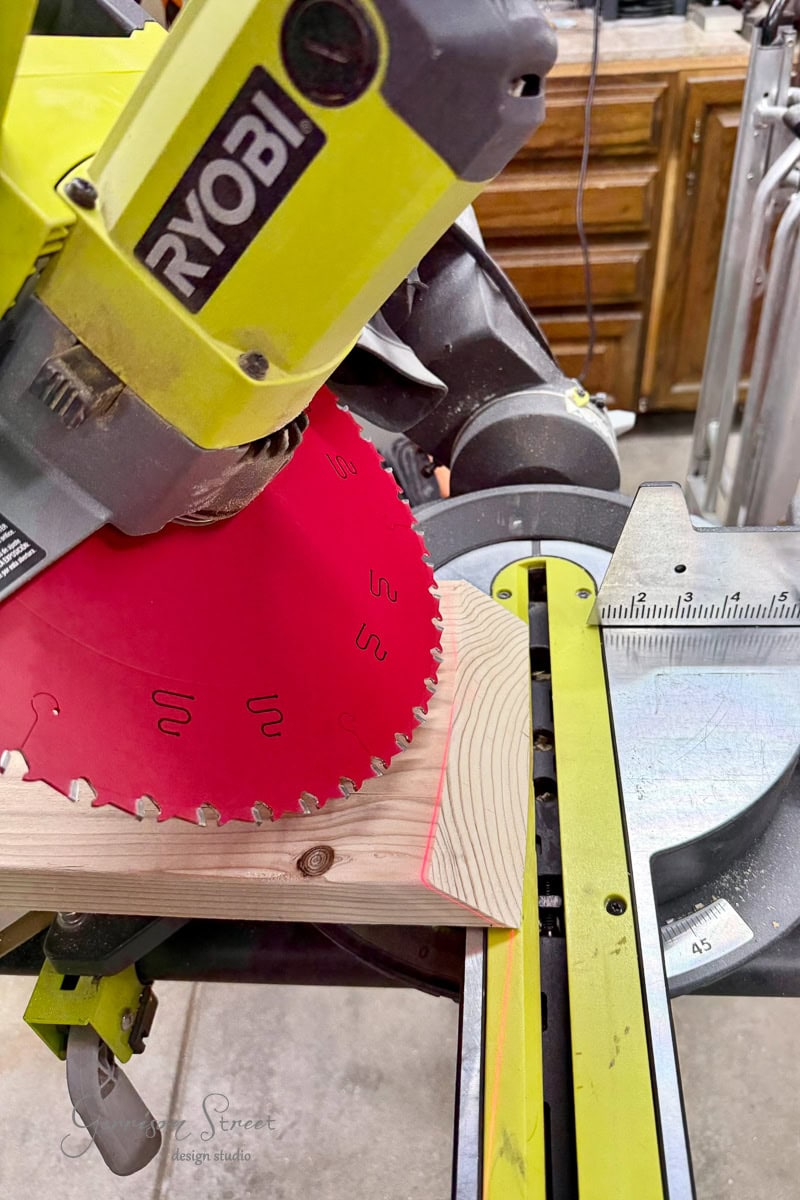 A Ryobi miter saw with a red blade is positioned over a piece of wood, ready to make a precise compound angled cut. The wood is marked with a red guideline. Workshop cabinets and tools are visible in the background.