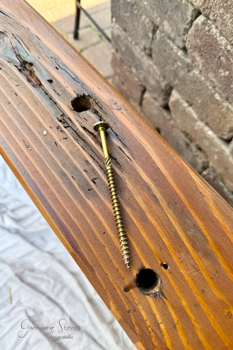 A gold wood screw lies on a wooden 6x6 near two uneven holes, with bricks in the background and a white cloth partially visible below.