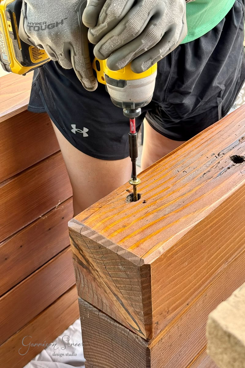 A girl wearing gloves and shorts uses a power drill to drive a screw into a wooden 6x6, with multiple 6x6s visible in the background.