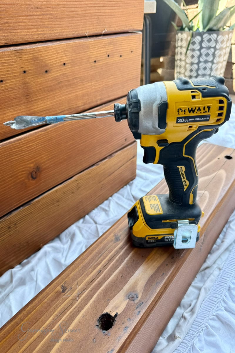 A yellow and black DeWalt power drill with a spade bit is resting on a wooden 6x6. There is a hole drilled in the wood, with more stacked 6x6s and a plant in the background.