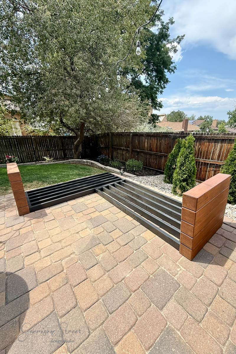 A partially constructed outdoor bench with wooden ends and black slats stands on a paved patio. A wooden fence, trees, and shrubs line the backyard under a blue sky with some clouds.