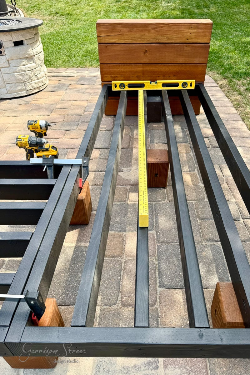 A wooden 2x4 frame under construction sits on a patio. A level tool rests across the slats, and a drill and clamps are visible. The background shows grass and a round stone fire pit.