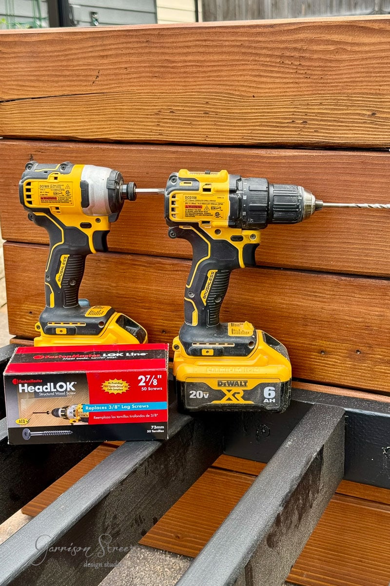 Two yellow DeWalt power drills rest on a wooden surface near a box of HeadLOK screws. A wooden fence is in the background, and the drills are placed on a black 2x4 frame.