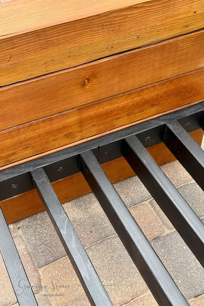 Black 2x4 support frames are attached to a wooden bench with a warm brown finish, sitting on a patio made of tan and brown stone pavers.