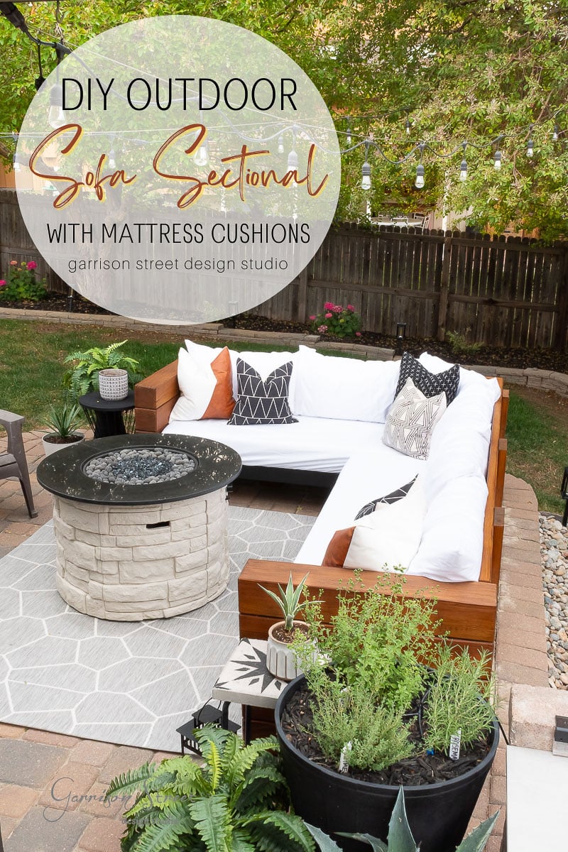 Outdoor patio with L-shaped wooden sofa featuring white mattress cushions, brown and patterned pillows, a round stone fire pit, potted plants, overhead string lights, and a large rug on the paver floor. Trees and a fence in the background.
