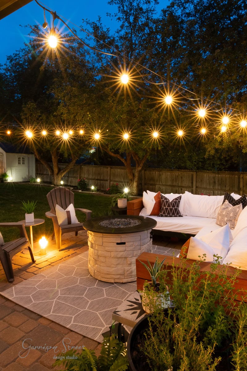 Cozy backyard patio at dusk with string lights overhead, a circular fire pit, Adirondack chairs, and a cushioned sectional sofa. Trees and plants surround the area.