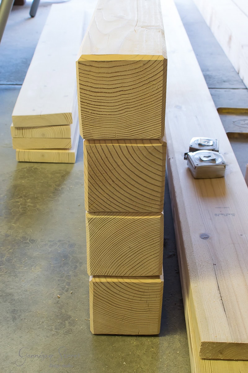 Four stacked wooden beams with visible wood grain patterns, lying on a workshop floor next to two tape measures and other pieces of lumber.