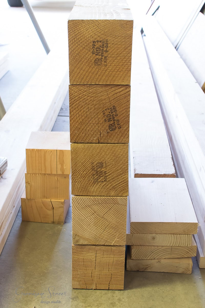 Stacked wooden beams and planks of various sizes are arranged vertically and horizontally in a workshop setting with additional loose boards in the background.