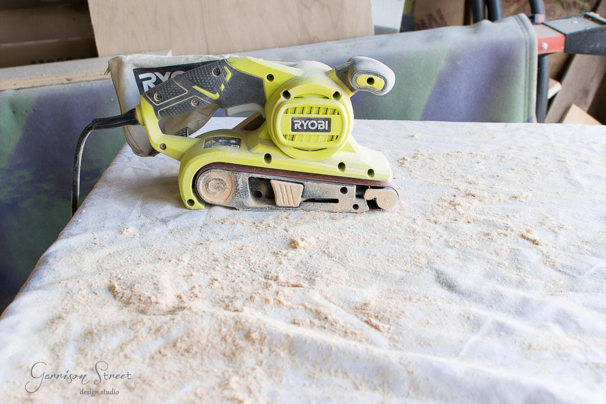 A yellow Ryobi belt sander sits on a cloth-covered workbench scattered with wood dust in a workshop setting.