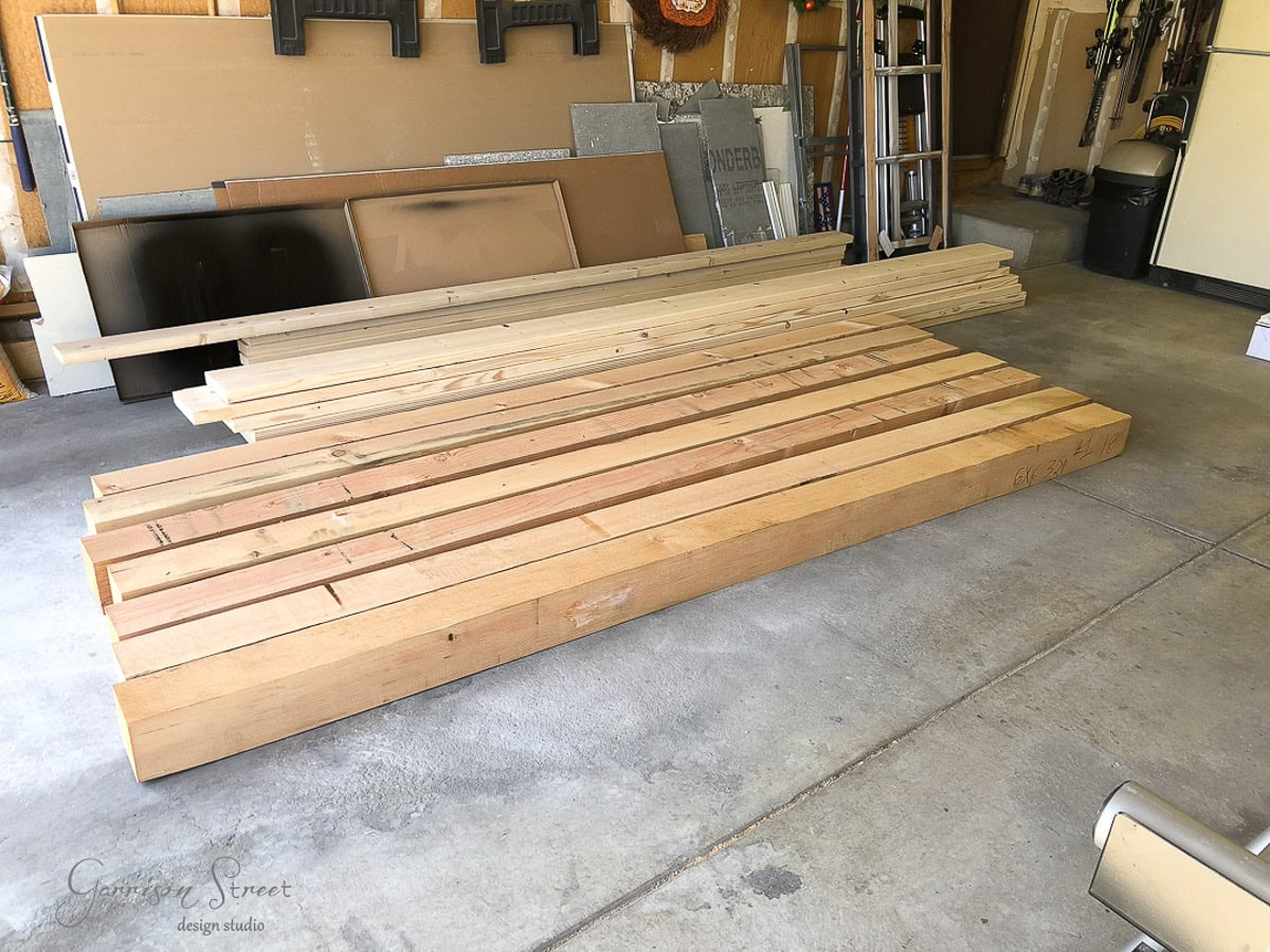 Several long wooden boards of varying sizes are stacked neatly on the floor of a garage, which has tools, metal sheets, and other materials organized against the walls.