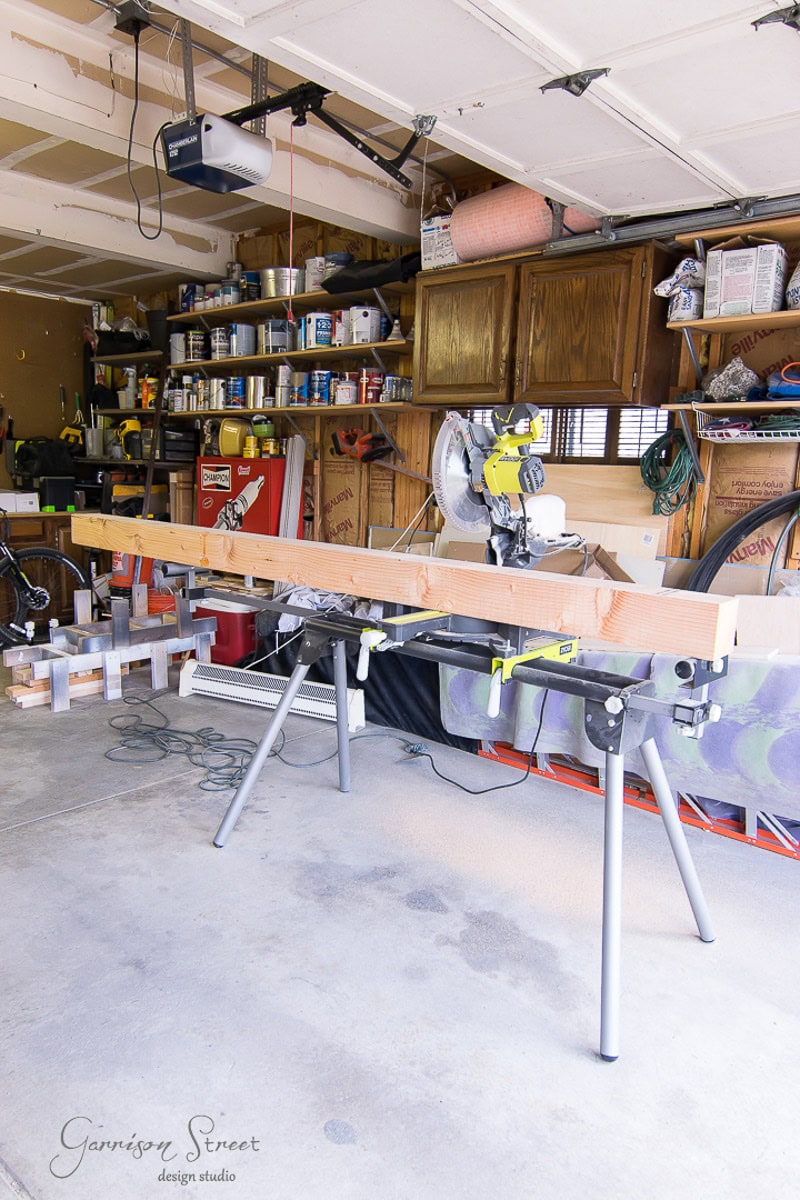 A garage workshop with a miter saw on a stand, a long wooden board ready for cutting, and shelves with tools, paint cans, and various supplies against the wall.