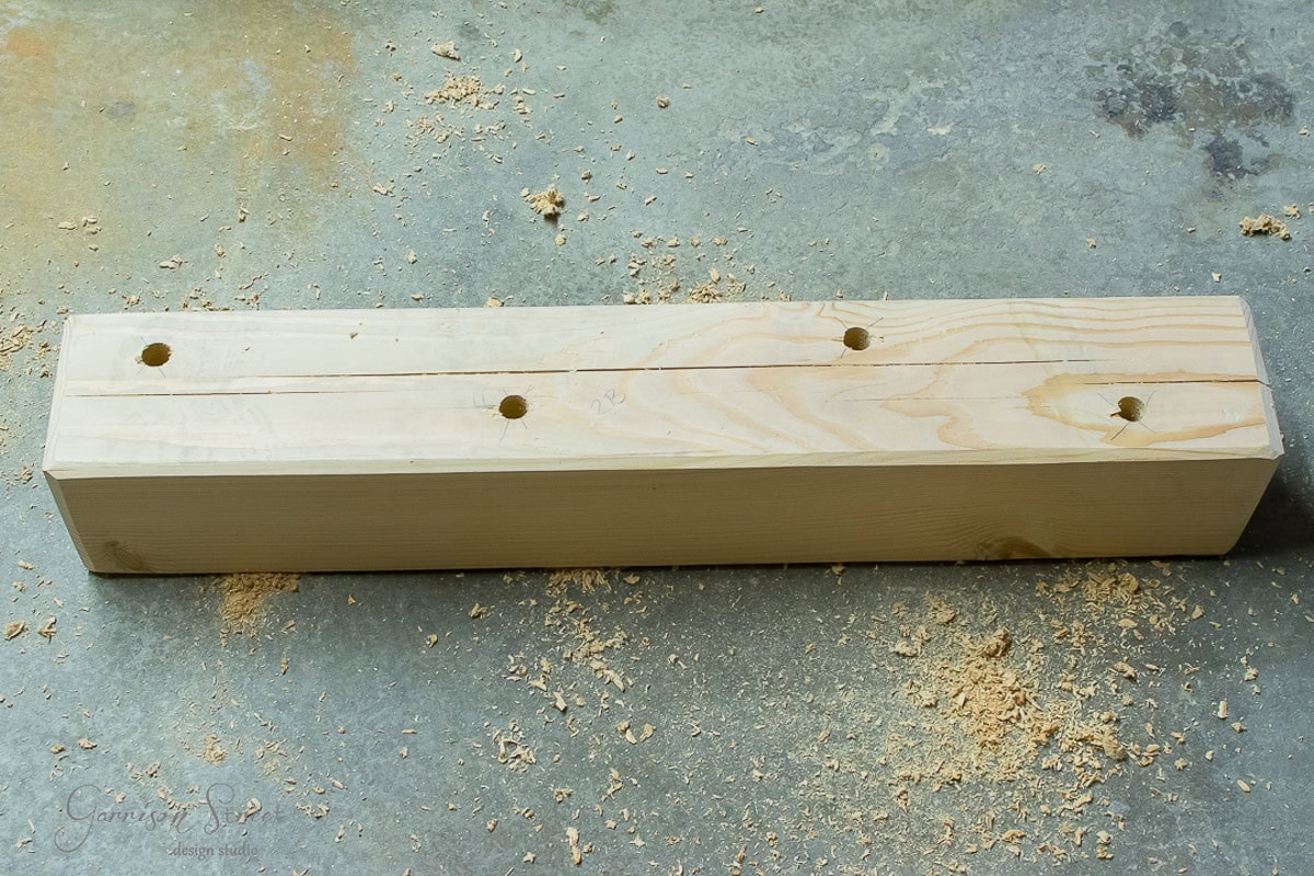 A wooden 6x6 with four evenly spaced holes drilled in it, lying on a concrete floor scattered with wood shavings.