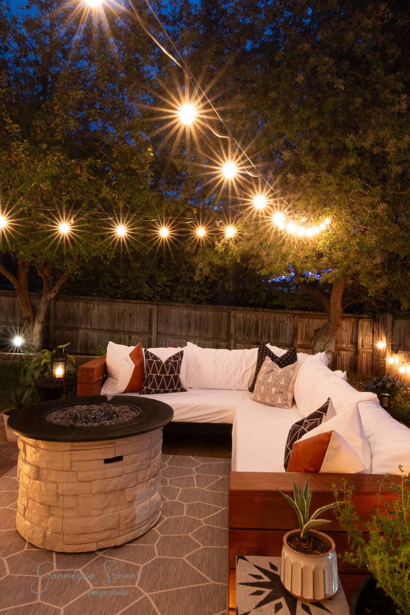 A cozy outdoor patio with string lights, a round stone fire pit, and a white cushioned sectional sofa, surrounded by trees and greenery at dusk. Pillows and potted plants add a welcoming touch.