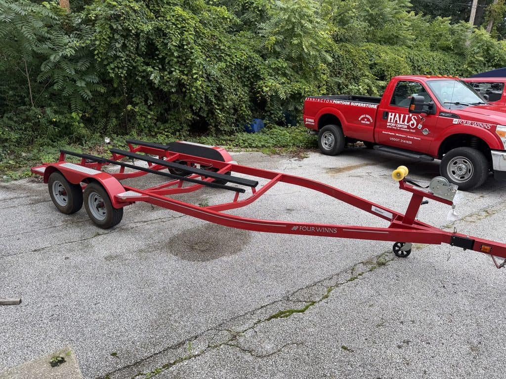 Red boat trailer parked on pavement with Hall's Sport Center truck nearby.