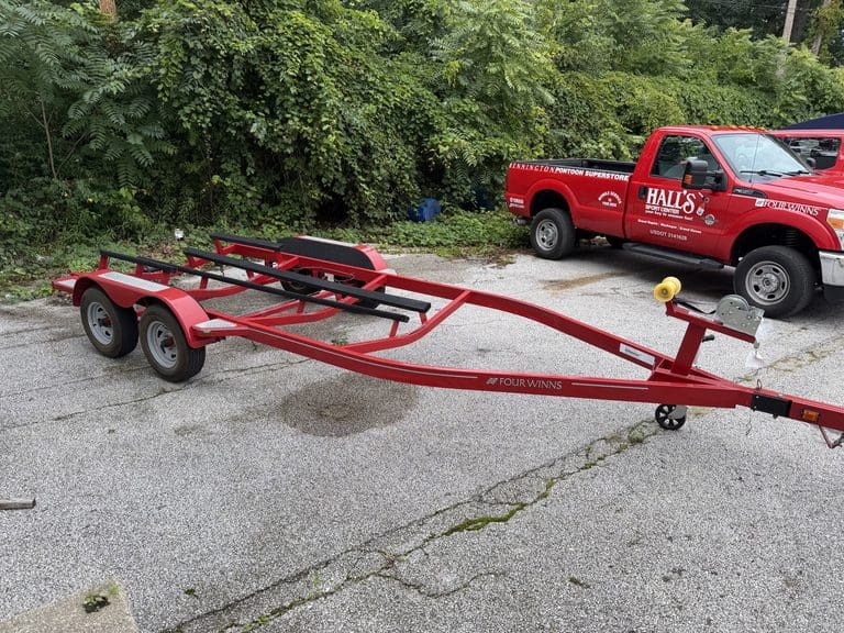 Red boat trailer parked on pavement with Hall's Sport Center truck nearby.