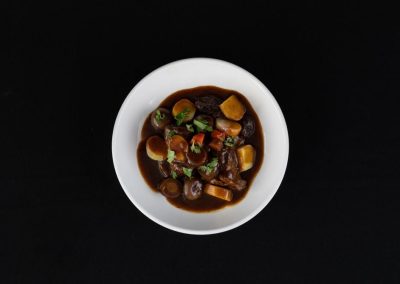 Delicious beef stew with vegetables served in a white bowl on black background.