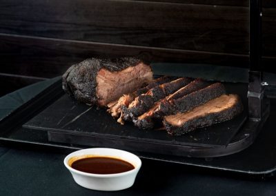 Smoked brisket slices on a black tray with BBQ sauce in a white bowl.