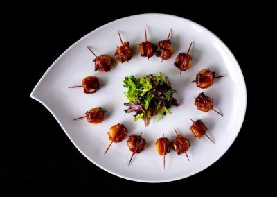 Bacon-wrapped skewers arranged in a circle on a white plate with greens in the center.