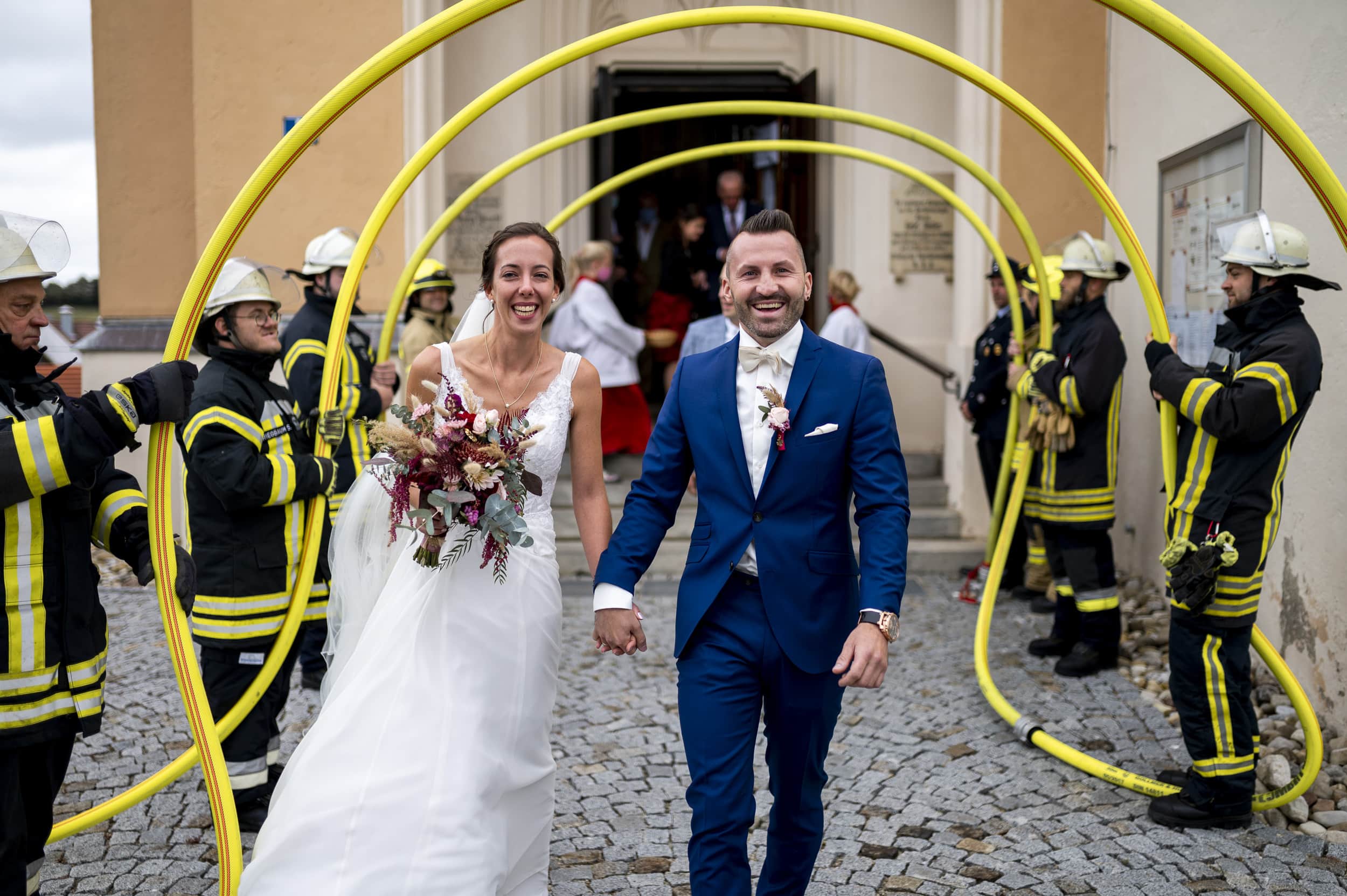 Feuerwehr steht bei Hochzeit vor Aindlinger Kirche Spalier und Brautpaar läuft durch Bogen aus Feuerwehrschläuchen