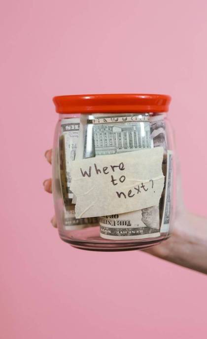 A hand holding a glass jar filled with dollar bills labeled 'Where to next?' against a pink background, symbolizing savings and future planning.