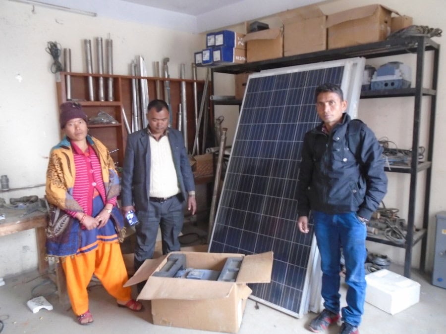 Three individuals, a woman and two men, standing next to a a number of large solar panels leaning against a wall.