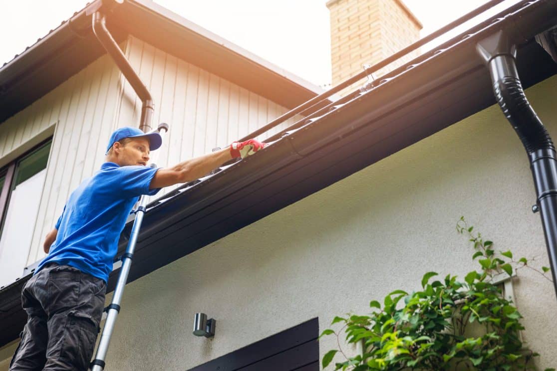 Worker cleaning gutters on residential house with ladder and safety gear.