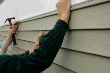 Worker installing vinyl siding on house exterior with hammer and nails.