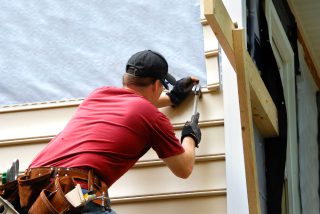 Worker installing siding on a house with tools and safety gear.