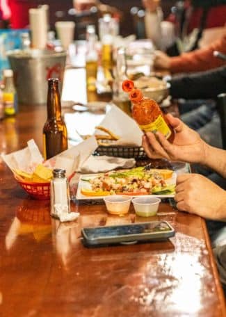 Person pouring hot sauce on a plate of food at a busy restaurant table.