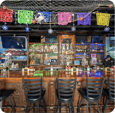 Colorful papel picado decorations over a bar with bottles and stools.