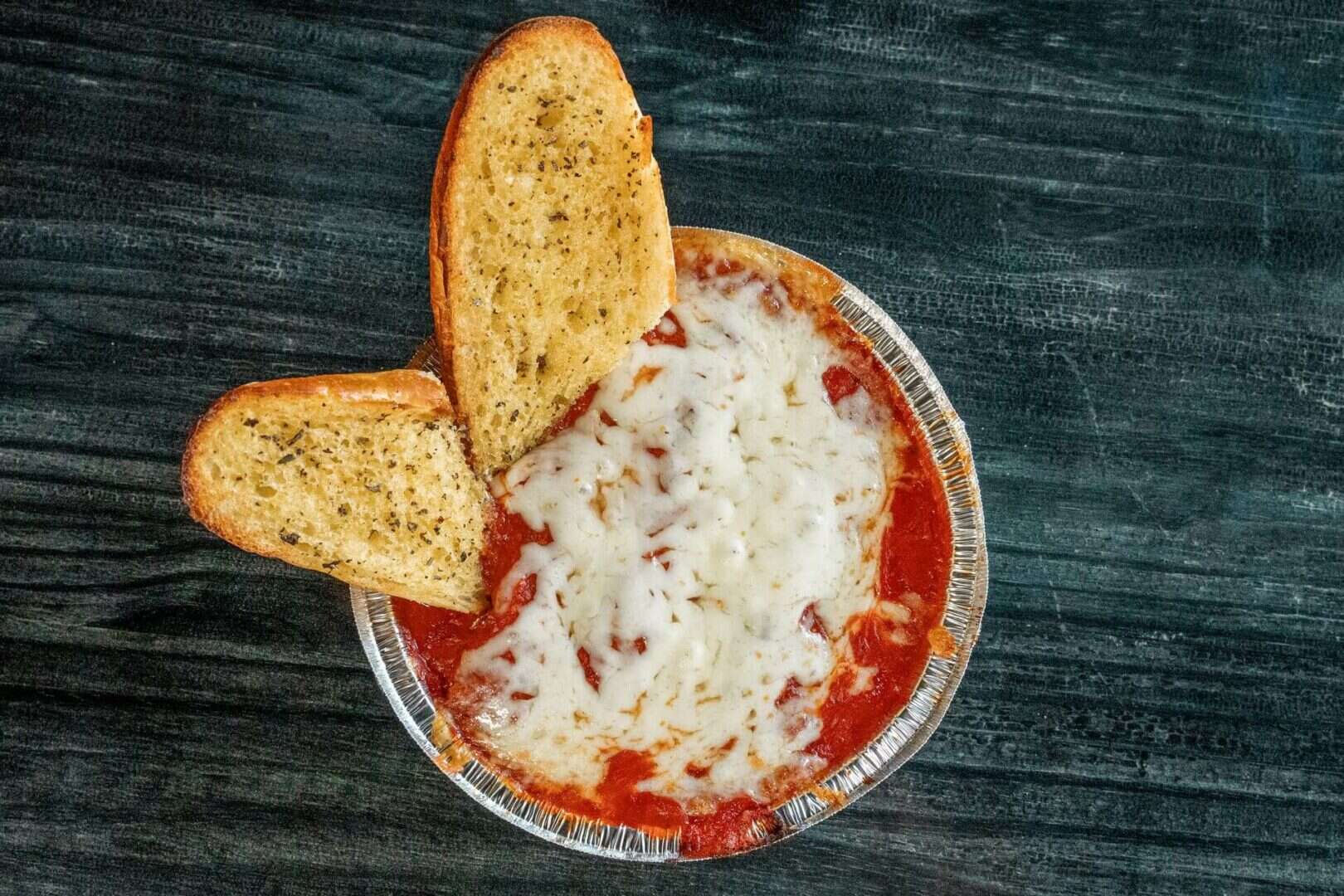 Sicilian pizza with melted cheese and garlic bread slices on dark wood table.