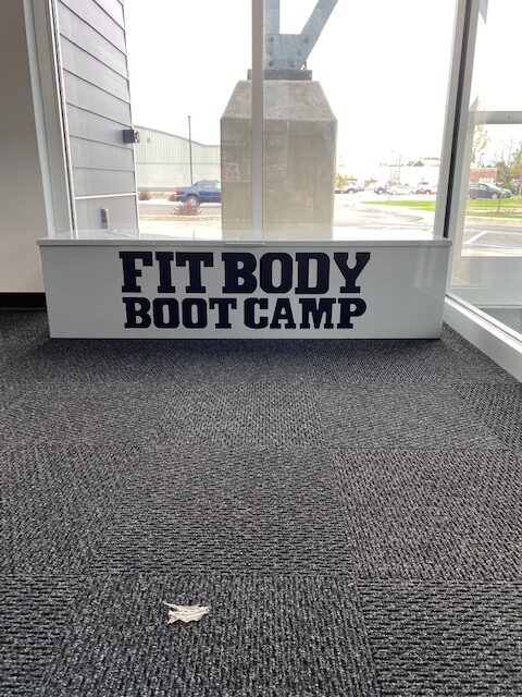 Large white sign with black text reading 'Fit Body Boot Camp' near window.