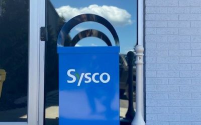Sysco sign and mailbox outside building with glass door and white brick wall.