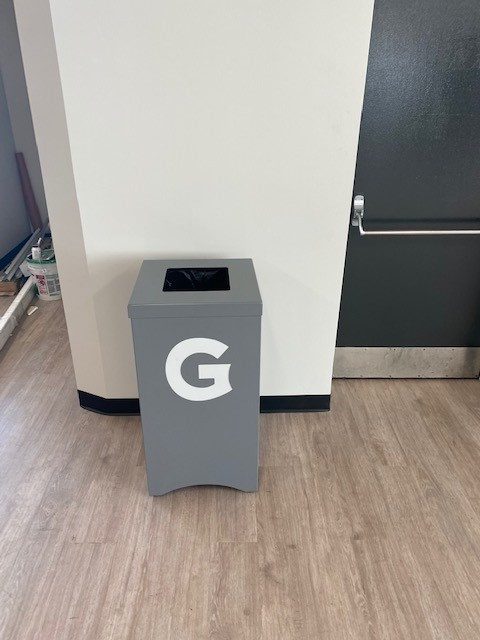 Gray trash bin with a white G logo in a commercial space.