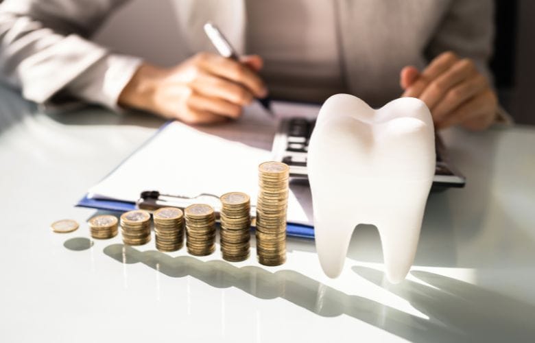 Stapel von Münzen vor einem Zahnmodell, im Hintergrund eine Person mit Taschenrechner – Symbolbild für die Finanzierung und Kostenplanung in einer Zahnarztpraxis.