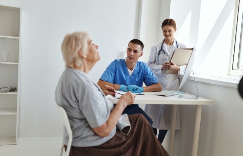 Ein Pfleger und eine Ärztin im aufmerksamen Gespräch mit einer älteren Patientin am Schreibtisch einer hellen Arztpraxis.