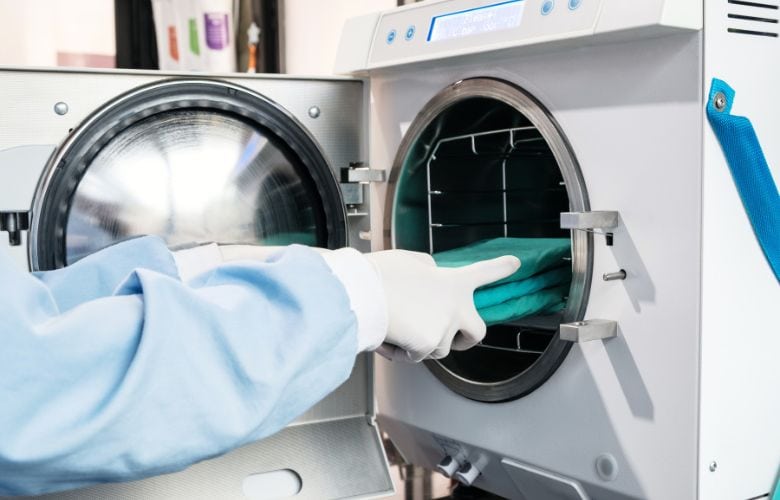Instrumentenaufbereitung in einem Sterilisationsraum mit moderner Ausrüstung Person in medizinischer Kleidung belädt einen offenen Autoklav in einer Arztpraxis mit grünen OP-Textilien.