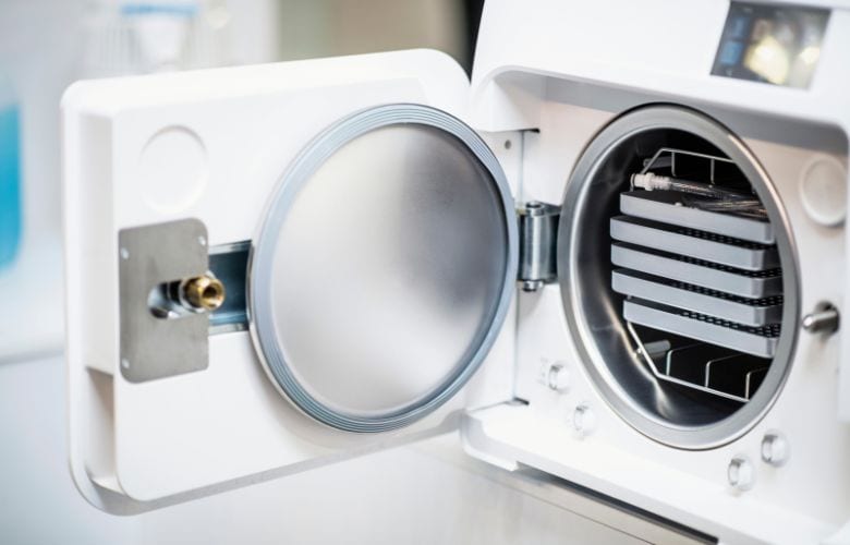 Geöffneter weißer Autoklav mit mehreren silbernen Einschub-Trays in der Sterilisationskammer.