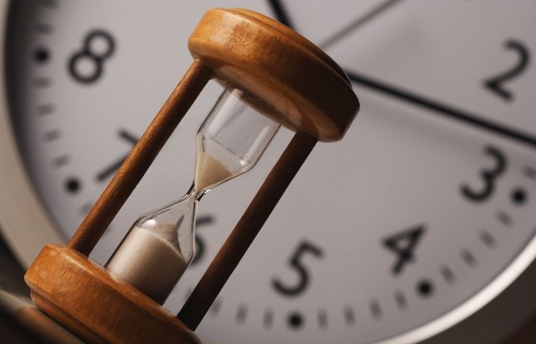 Eine hölzerne Sanduhr im Vordergrund vor dem unscharfen Ziffernblatt einer Wanduhr für effizientes Zeitmanagement.
