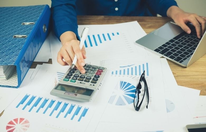 Mitarbeiter einer privatärztlichen Verrechnungsstelle arbeitet mit Taschenrechner, Stift und Laptop an der Auswertung von Finanzdiagrammen und Dokumenten am Schreibtisch.