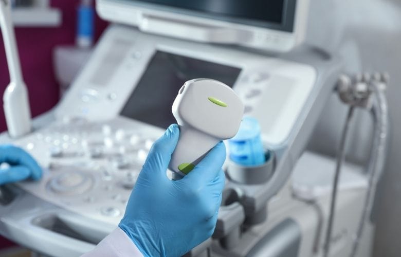 Hand in blauem Handschuh hält eine Ultraschallsonde vor einem medizinischen Ultraschallgerät und Gelbehälter.