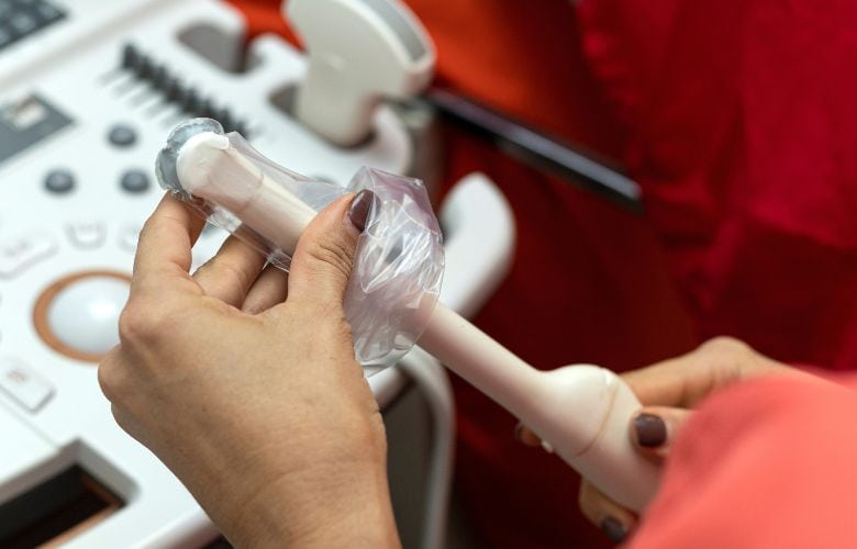 Hände ziehen eine Schutzhülle über eine endokavitäre Ultraschallsonde vor einer gynäkologischen Untersuchung.