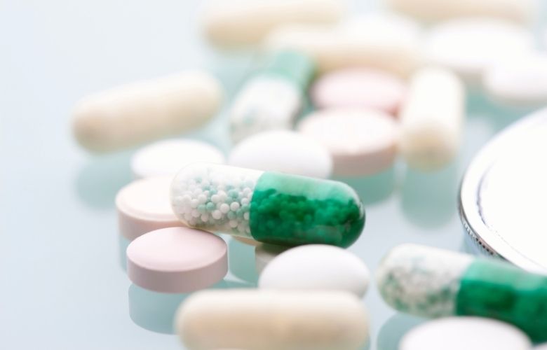 Verschiedene farbige Tabletten und eine grün-weiße Kapsel mit Mikropellets auf spiegelnder Glasfläche.