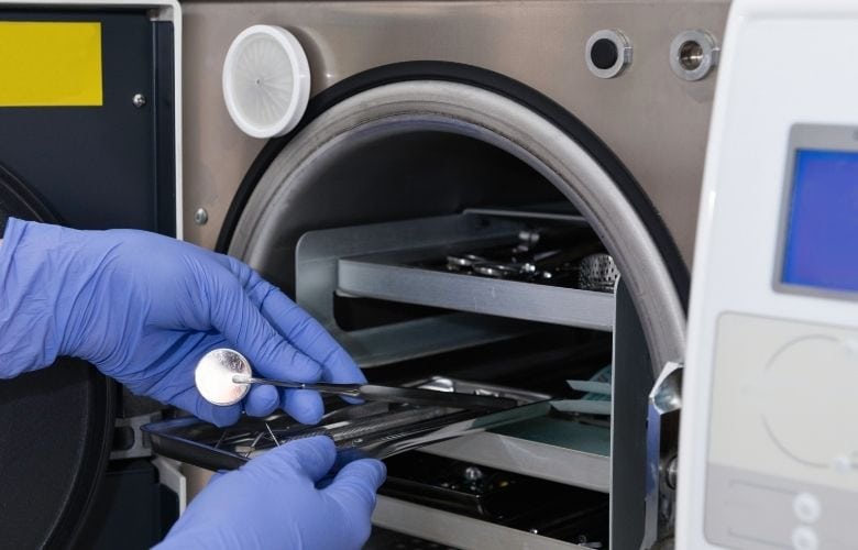 Person in blauen Handschuhen schiebt ein Metalltray mit Zahnarztinstrumenten zur Sterilisation in einen Autoklav.