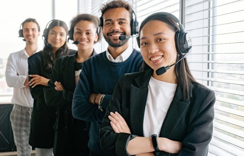 Fünf lächelnde Callcenter-Mitarbeiter mit Headsets stehen hintereinander in einem hellen Büro.