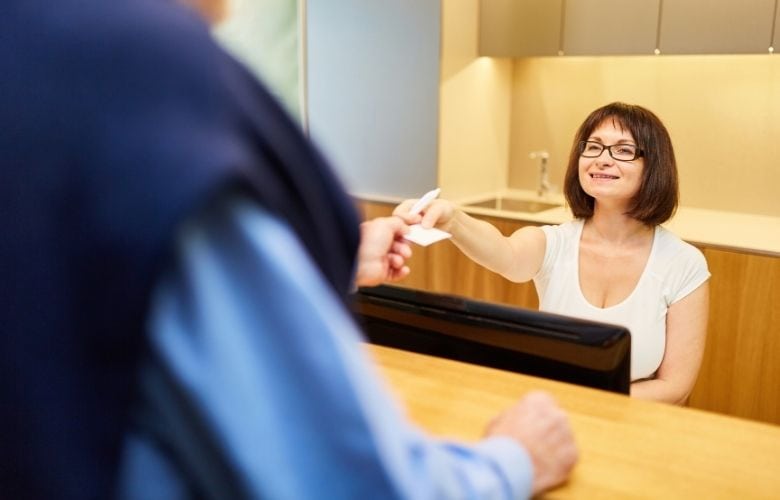 Freundliche Arzthelferin übergibt am Empfangstresen einer Praxis eine Karte an einen Patienten.