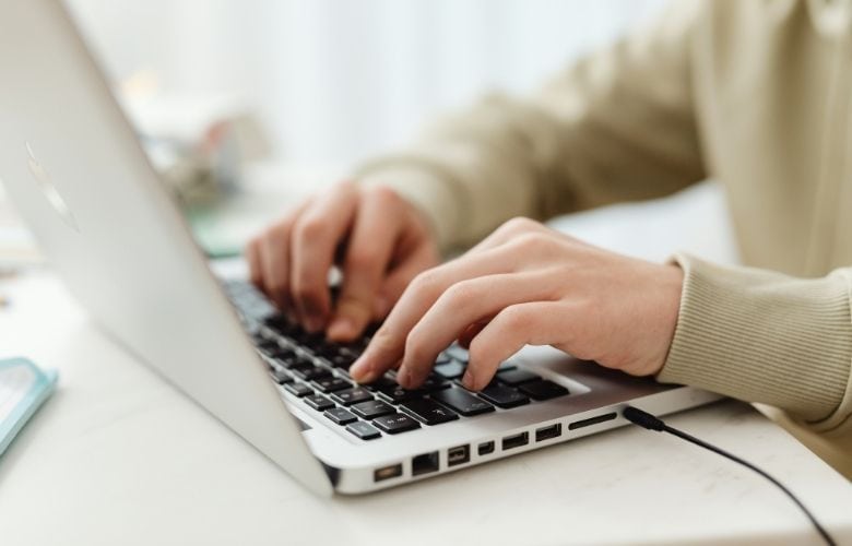 Zwei Hände tippen auf der Tastatur eines silbernen Laptops auf einem hellen Schreibtisch.